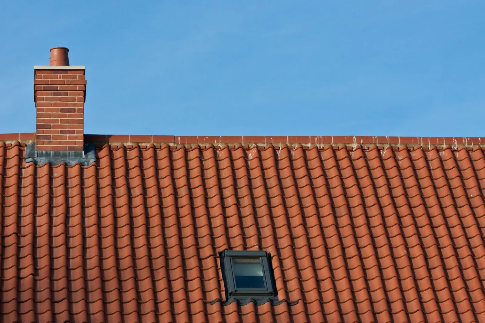 Rood pannendak met schoorsteen en dakraam tegen een blauwe lucht
